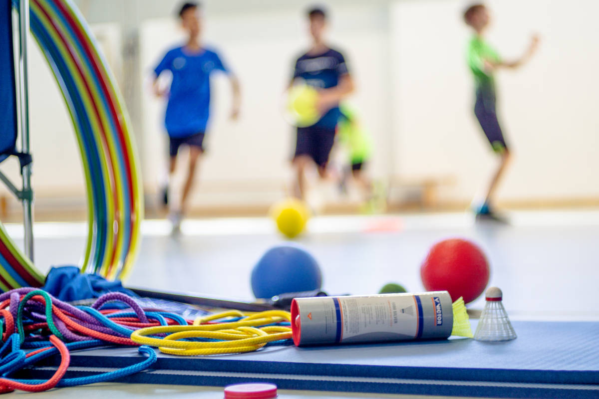 Im Vordergund sind verschiedene Sportutensilien zu sehen, während Kinder im Hintergrund in der Turnhalle Sport treiben