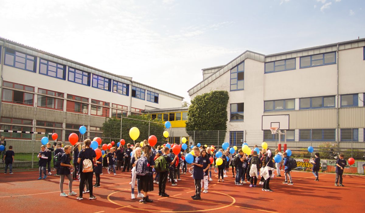 Schüler stehen auf dem Hartplatz und halten bunte Luftballons in der Hand