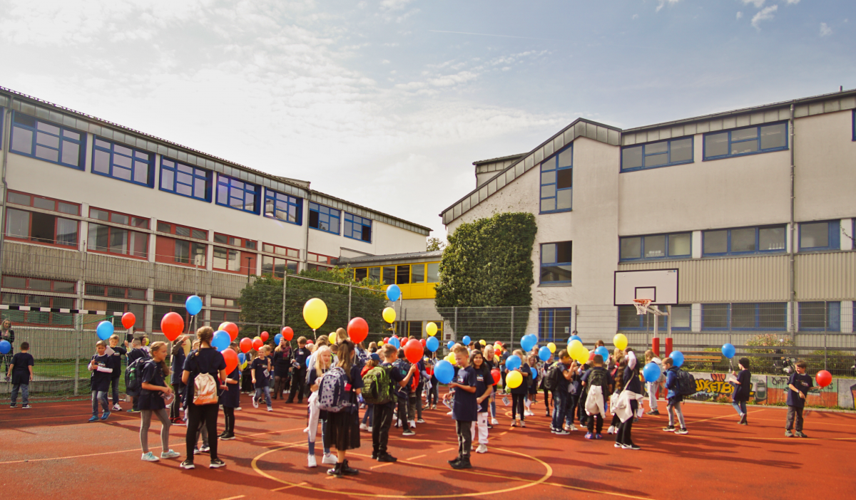 Schüler stehen auf dem Hartplatz und halten bunte Luftballons in der Hand