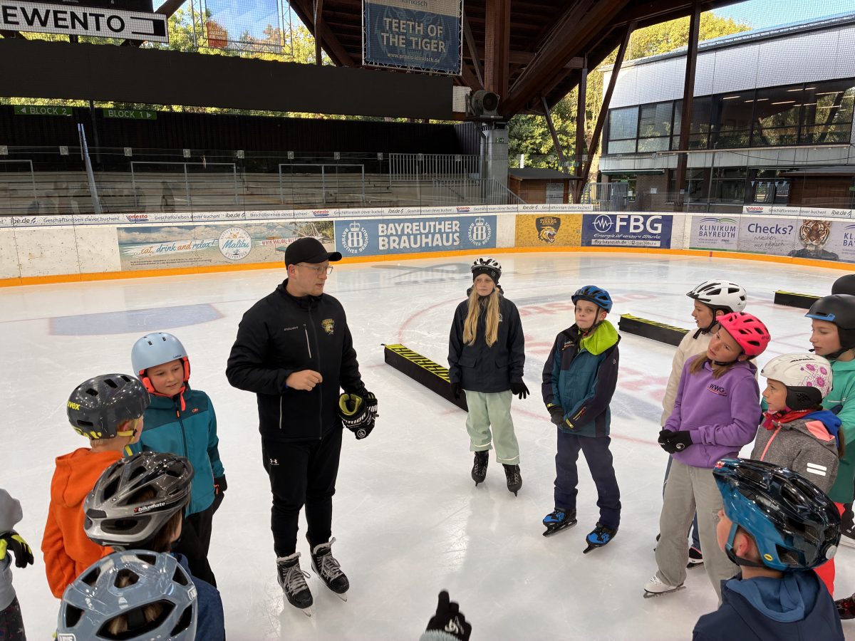 Ein Eishockeytrainer steht mit der Klasse auf dem Eis und hält einen Vortrag